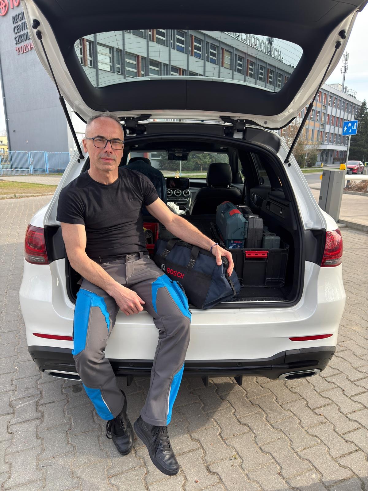 Mężczyzna w spodniach roboczych siedzi na bagażniku białego auta, obok torba Bosch i skrzynki narzędziowe. W tle budynek z napisem.