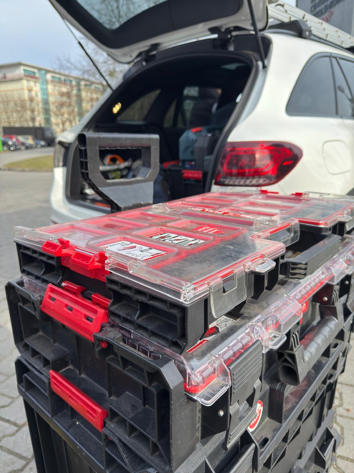 Zestaw skrzynek narzędziowych na tle otwartego bagażnika białego kombi z drabiną na dachu, sugerujący gotowość do pracy w terenie.