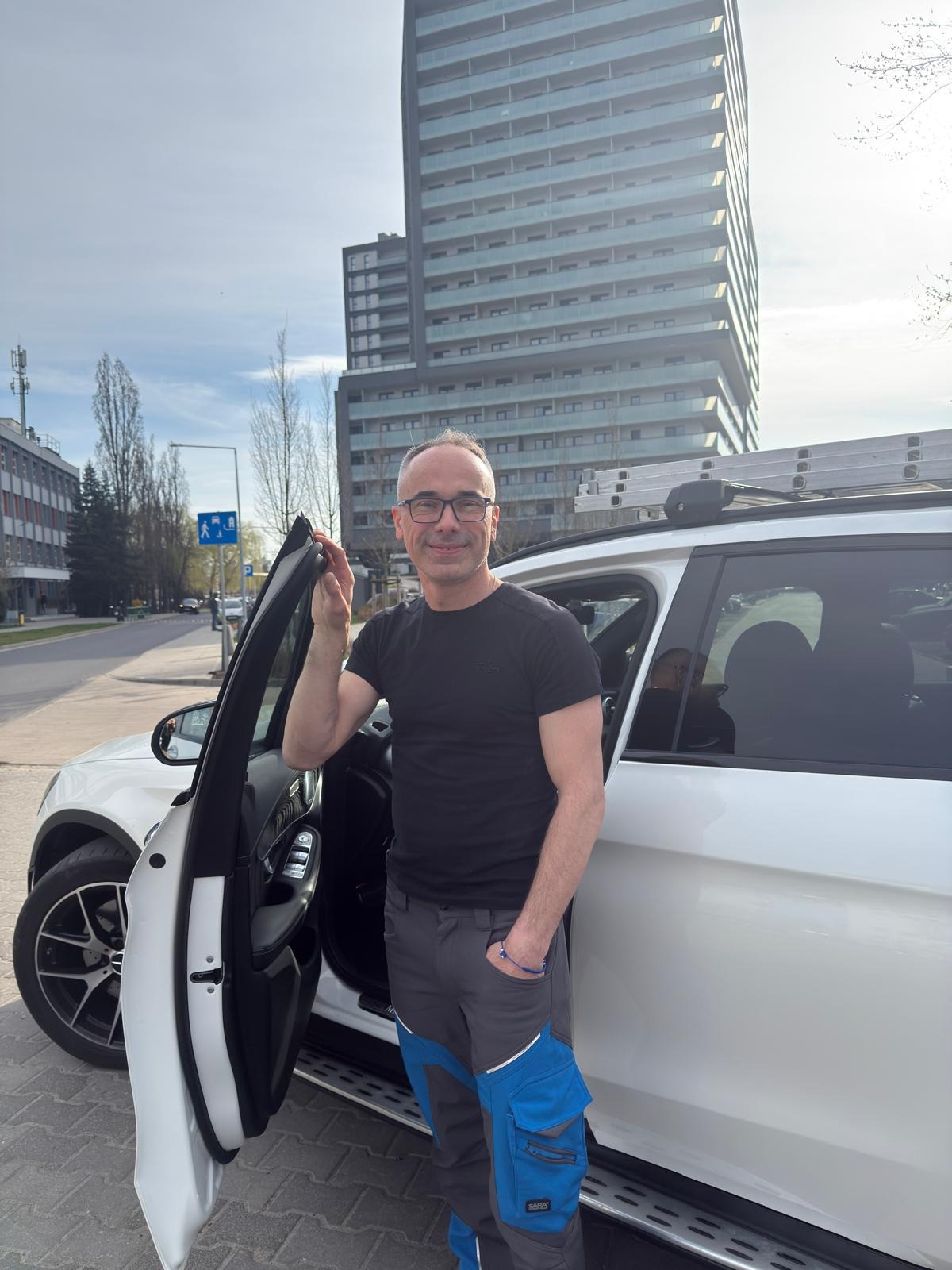 Uśmiechnięty mężczyzna w roboczych spodniach stoi przy białym samochodzie z drabiną na dachu, na tle miejskiego krajobrazu z wysokim budynkiem.