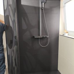 STONEFORT sp. z o.o - Nowoczesna kabina prysznicowa z deszczownicą i czarnymi płytkami. Widoczny fragment osoby montującej kabinę. Minimalistyczny design i precyzyjne wykończenie.