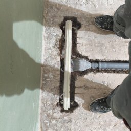 STONEFORT sp. z o.o - Montaż odpływu liniowego w podłodze łazienki, widok z góry na fragment instalacji i fragment ściany z płyt karton-gips. Widoczne buty montera.