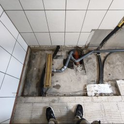 STONEFORT sp. z o.o - Ułożona instalacja odpływowa w łazience z widocznym odpływem liniowym, rurami PCV i kablami elektrycznymi przed wylaniem posadzki. Białe płytki na ścianach.