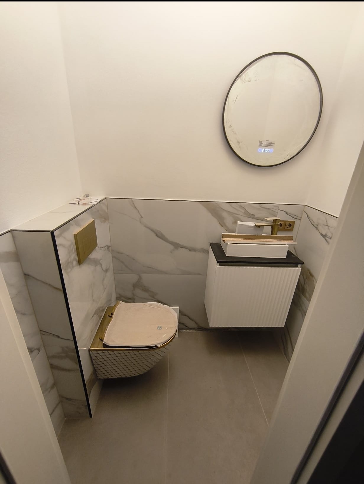 Elegancka toaleta z marmurowymi płytkami, złotymi akcentami i okrągłym lustrem z podświetleniem. Nowoczesna armatura i minimalistyczny design.