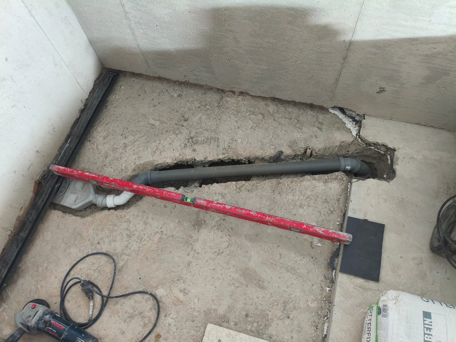 Szare rury kanalizacyjne w wykopanym bruździe w podłodze, poziomica, szlifierka kątowa i worek zaprawy w trakcie prac instalacyjnych w pomieszczeniu.