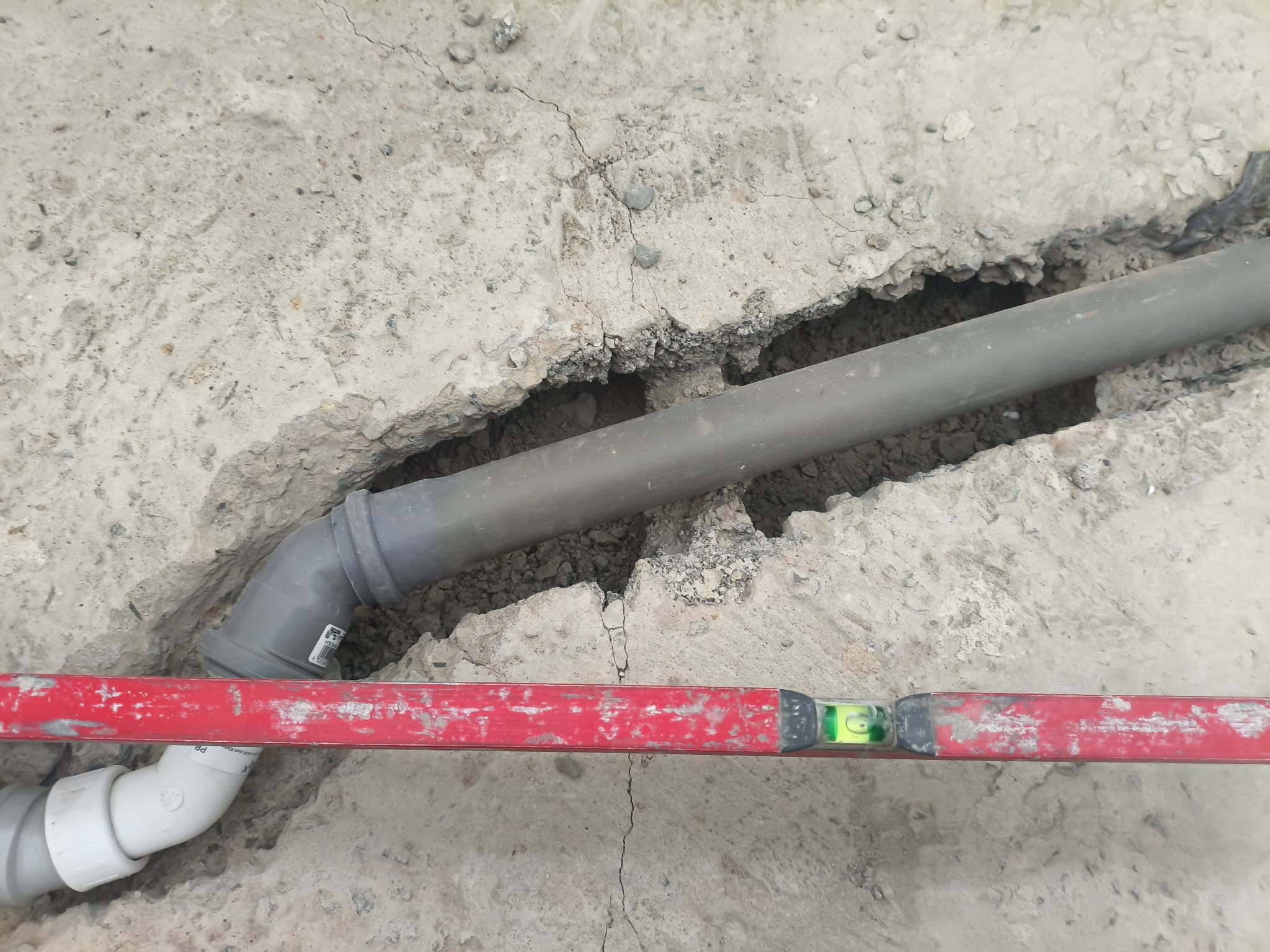 Szara rura kanalizacyjna ułożona w wyżłobieniu w betonowej posadzce, poziomowana czerwoną poziomicą. Widoczne złącza i fragmenty instalacji.