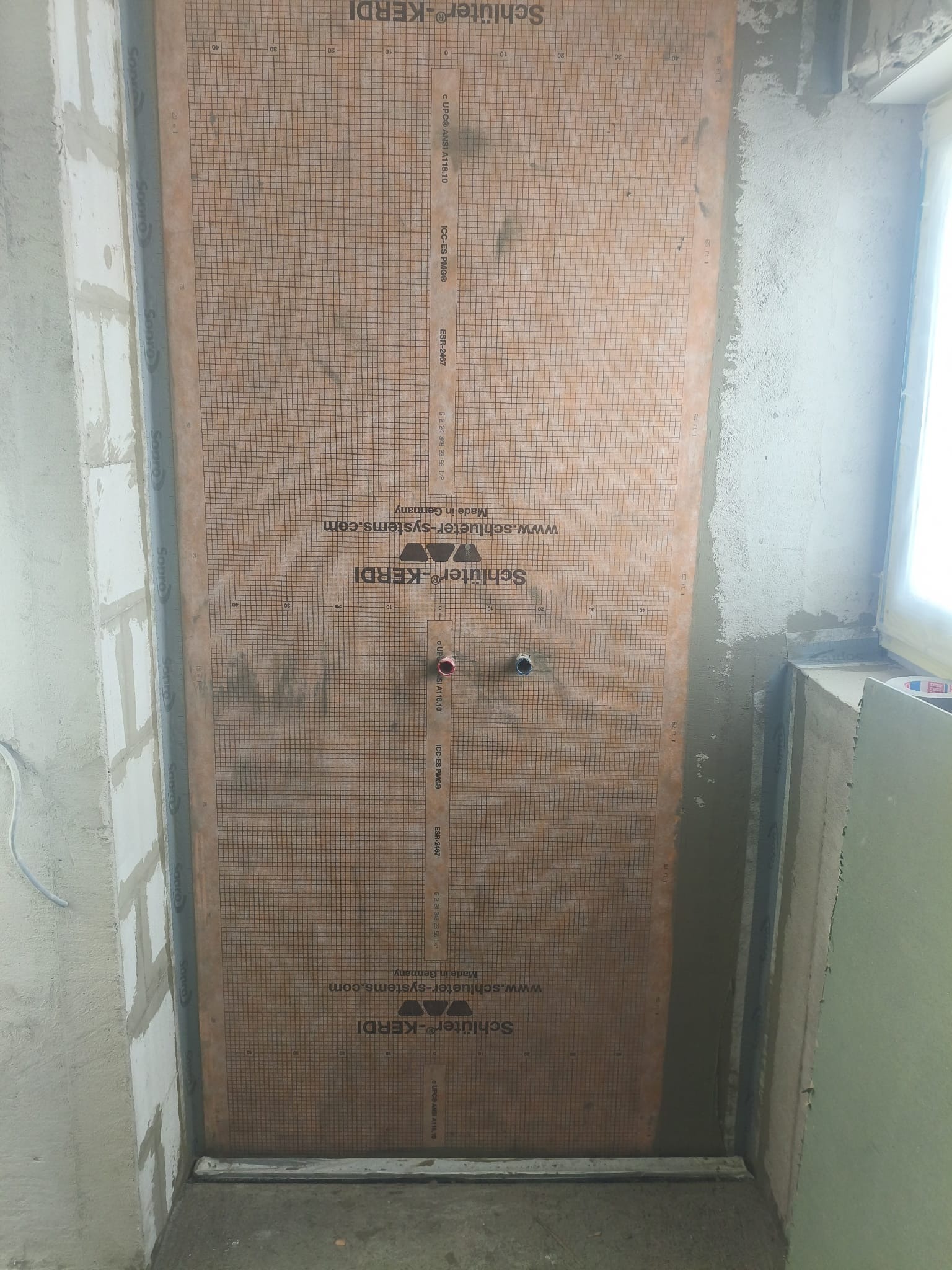 Przygotowana ściana z matą uszczelniającą Kerdi w łazience przed położeniem płytek. Widoczne wyprowadzenia instalacji wodnej i fragment okna.