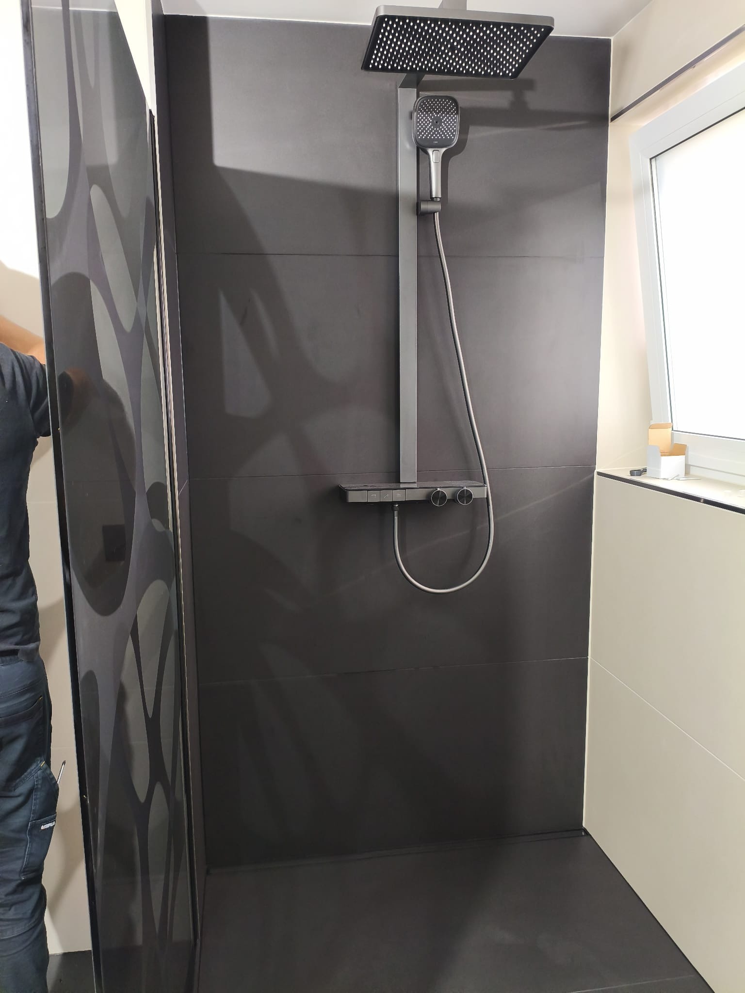 Nowoczesna kabina prysznicowa z deszczownicą i czarnymi płytkami. Widoczny fragment osoby montującej kabinę. Minimalistyczny design i precyzyjne wykończenie.