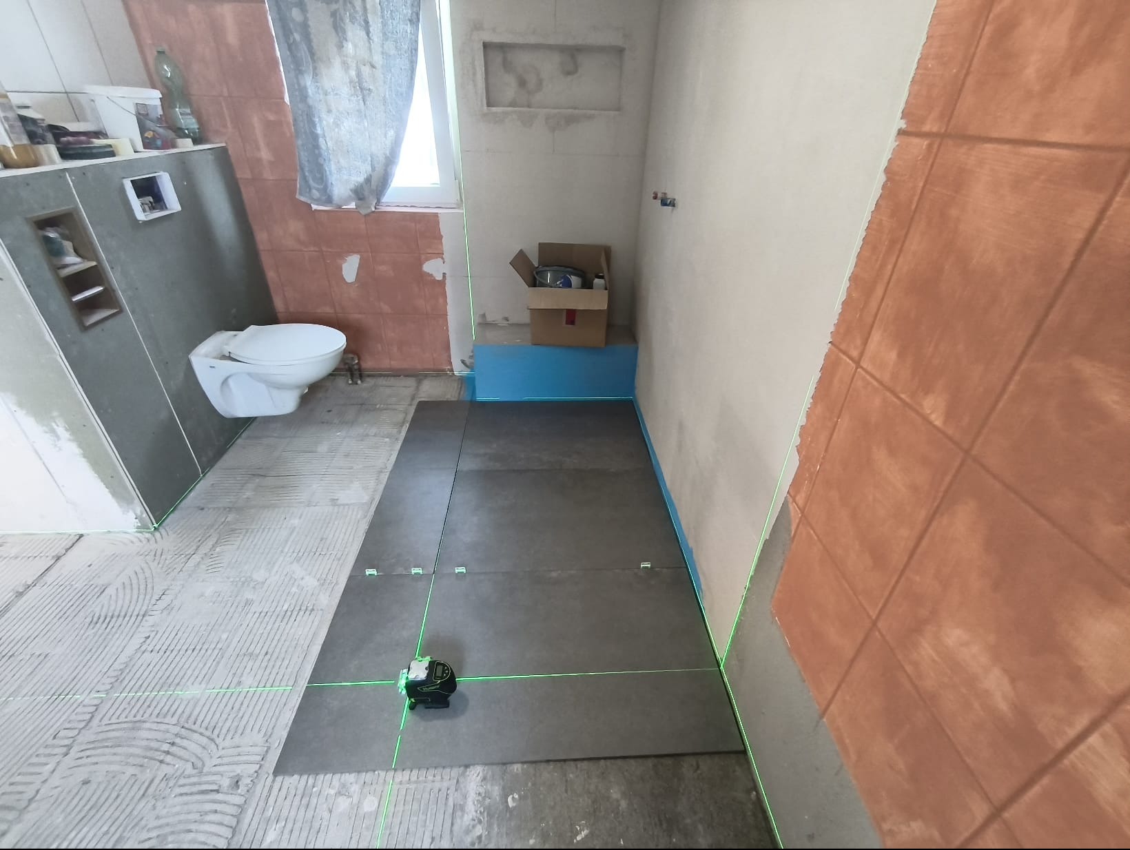 Wnętrze łazienki w trakcie prac. Częściowo ułożone szare płytki podłogowe, poziomica laserowa, widoczne płytki ścienne i przygotowane ściany. Etap prac wykończeniowych.