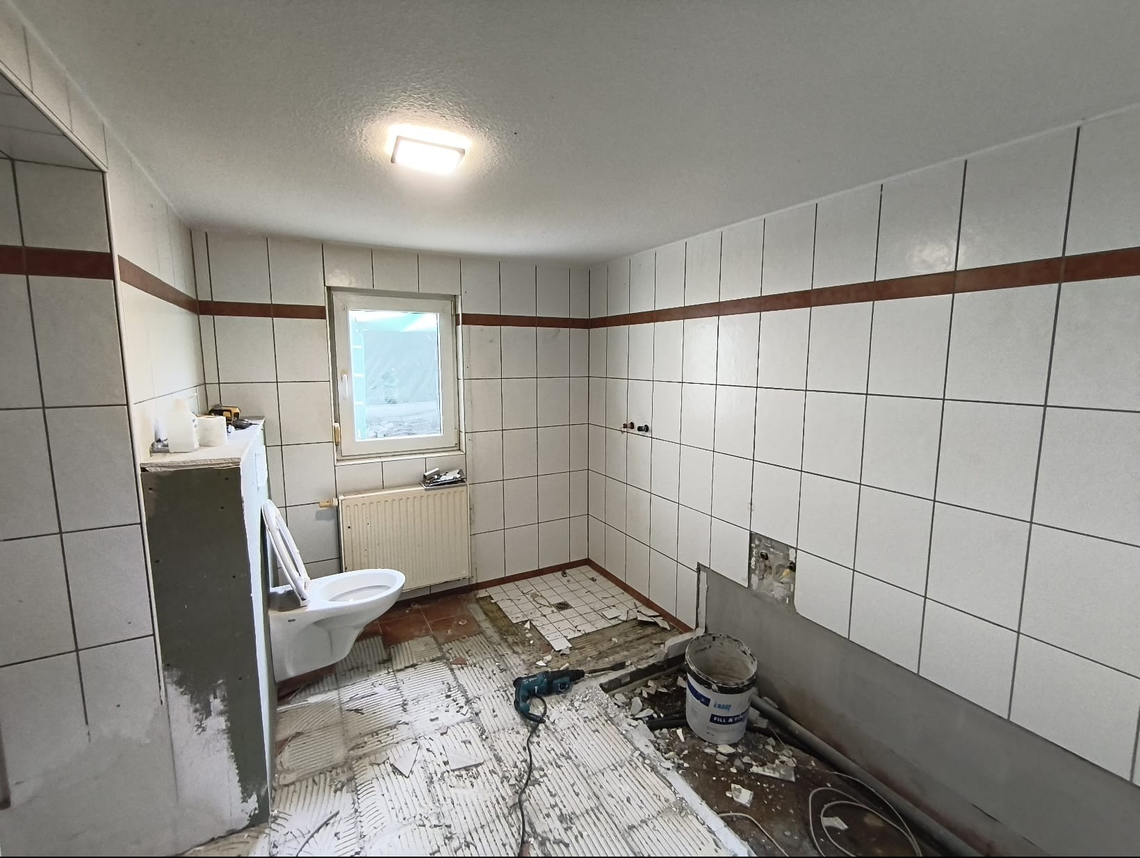 Łazienka w trakcie demontażu płytek. Widoczne narzędzia, odspojone płytki podłogowe, częściowo zdemontowana toaleta. Ściany z białymi płytkami i brązowym pasem.