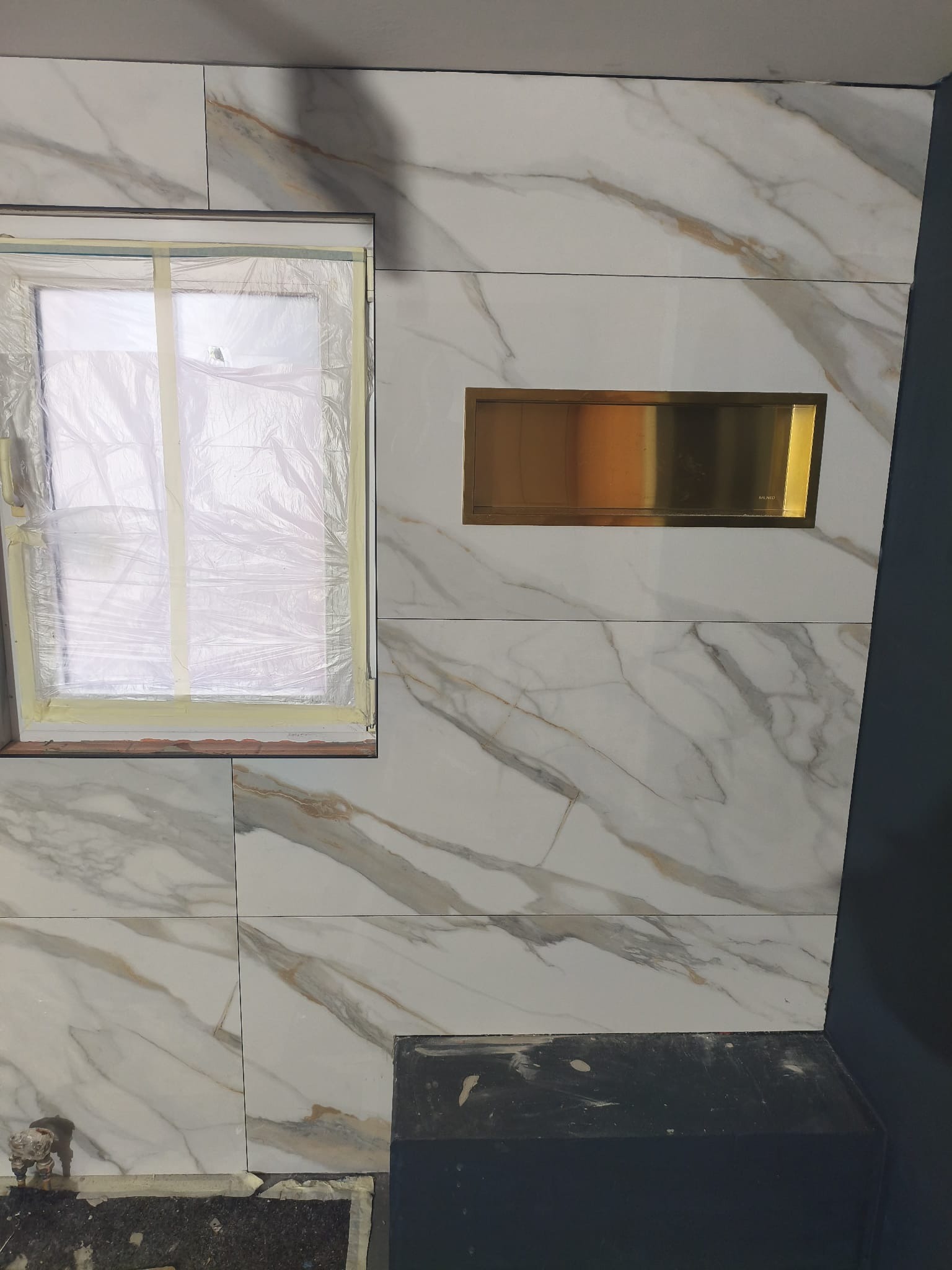 Ściana z płytek imitujących marmur z wnęką w kolorze złotym. Okno zabezpieczone folią malarską. Fragment podłogi w trakcie prac wykończeniowych.