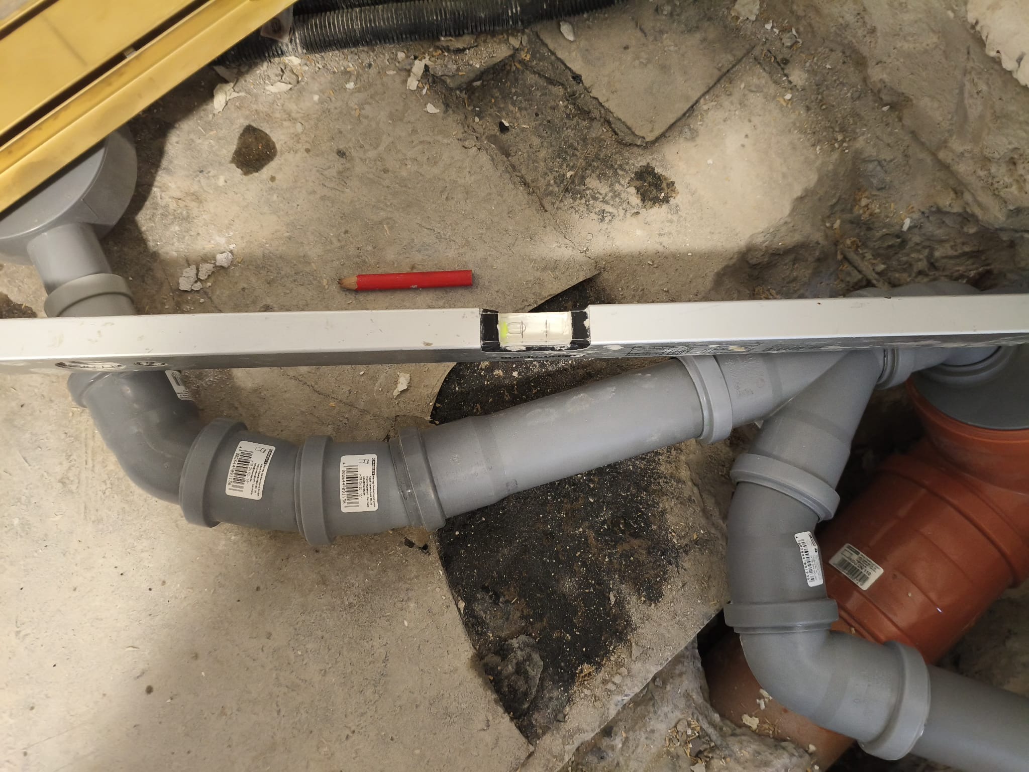 Szare rury kanalizacyjne ułożone w wykopie, poziomowane poziomicą. Widoczny fragment odpływu liniowego i czerwona kredka stolarska. Prace instalacyjne w trakcie remontu.