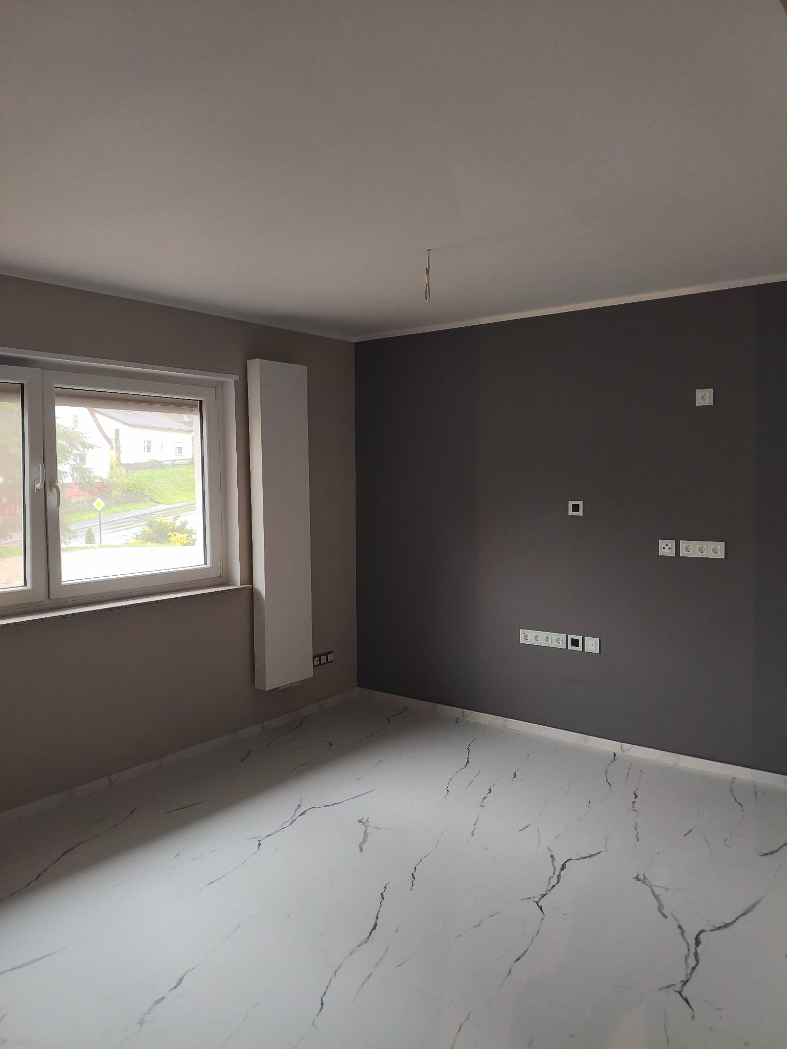 Wnętrze po odnowieniu: biała marmurowa podłoga, szare ściany z gniazdkami, biały grzejnik pionowy i okno z widokiem na zieleń. Minimalistyczny design.