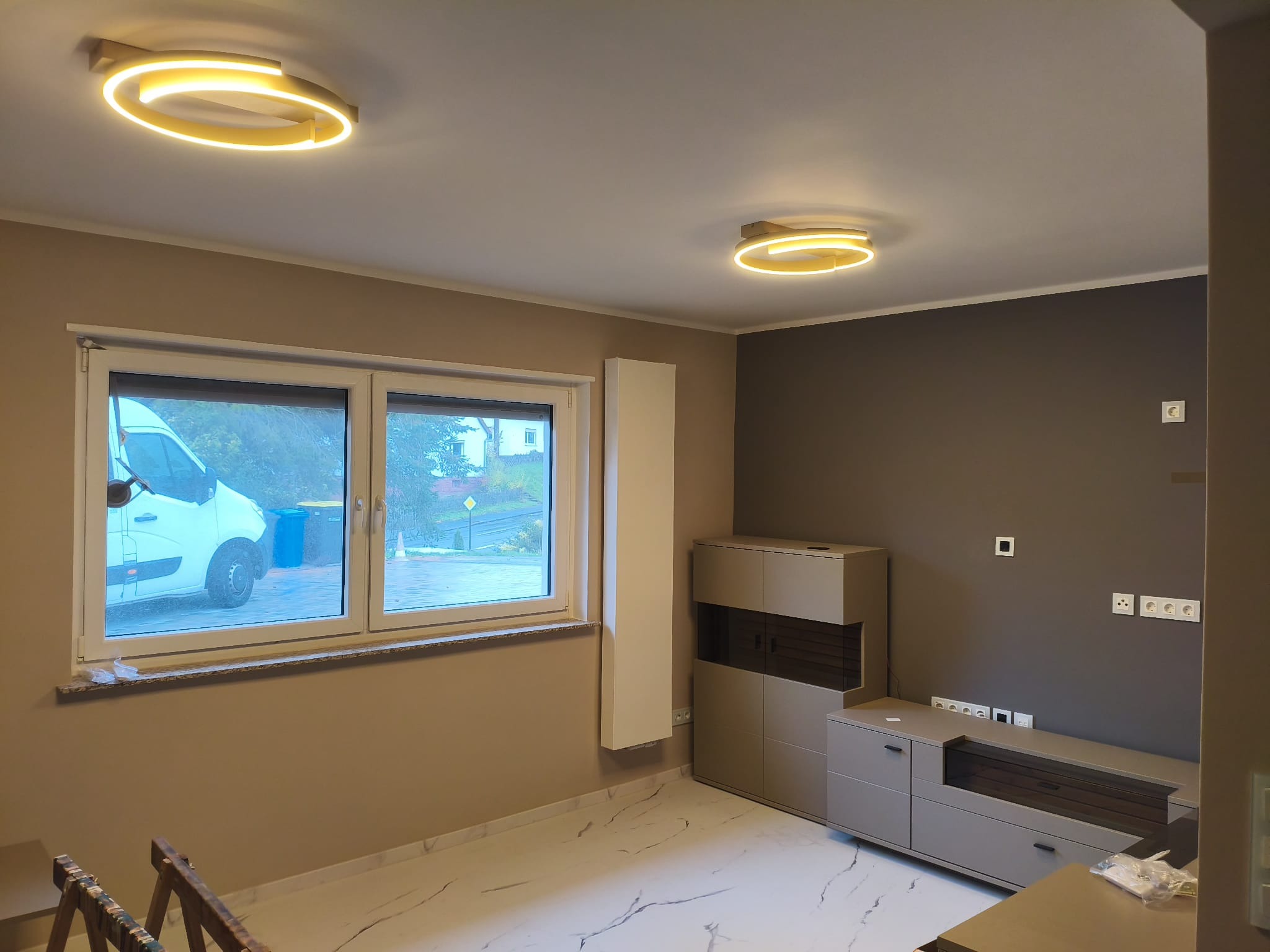 Pokój po remoncie: świeżo pomalowane ściany w odcieniach beżu i szarości, nowoczesne oświetlenie sufitowe, marmurowe płytki podłogowe, widok na zewnątrz przez okno.