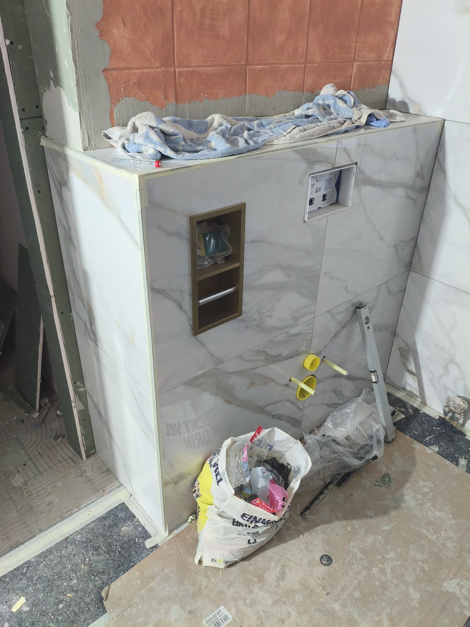 Wnętrze łazienki w trakcie wykańczania: obudowa WC z marmurowymi płytkami, wnęka z półkami, widoczne narzędzia i materiały budowlane. Etap prac wykończeniowych.