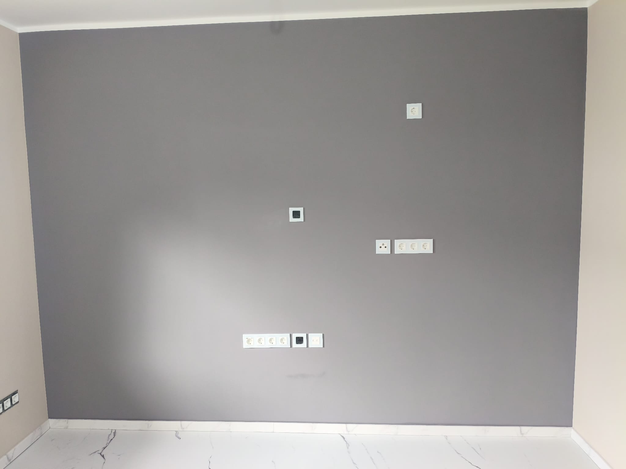 Ściana pomalowana na szaro z gniazdkami elektrycznymi i wykończoną podłogą z białego marmuru. Widoczne listwy przypodłogowe i fragment sąsiedniej ściany w odcieniu beżu.
