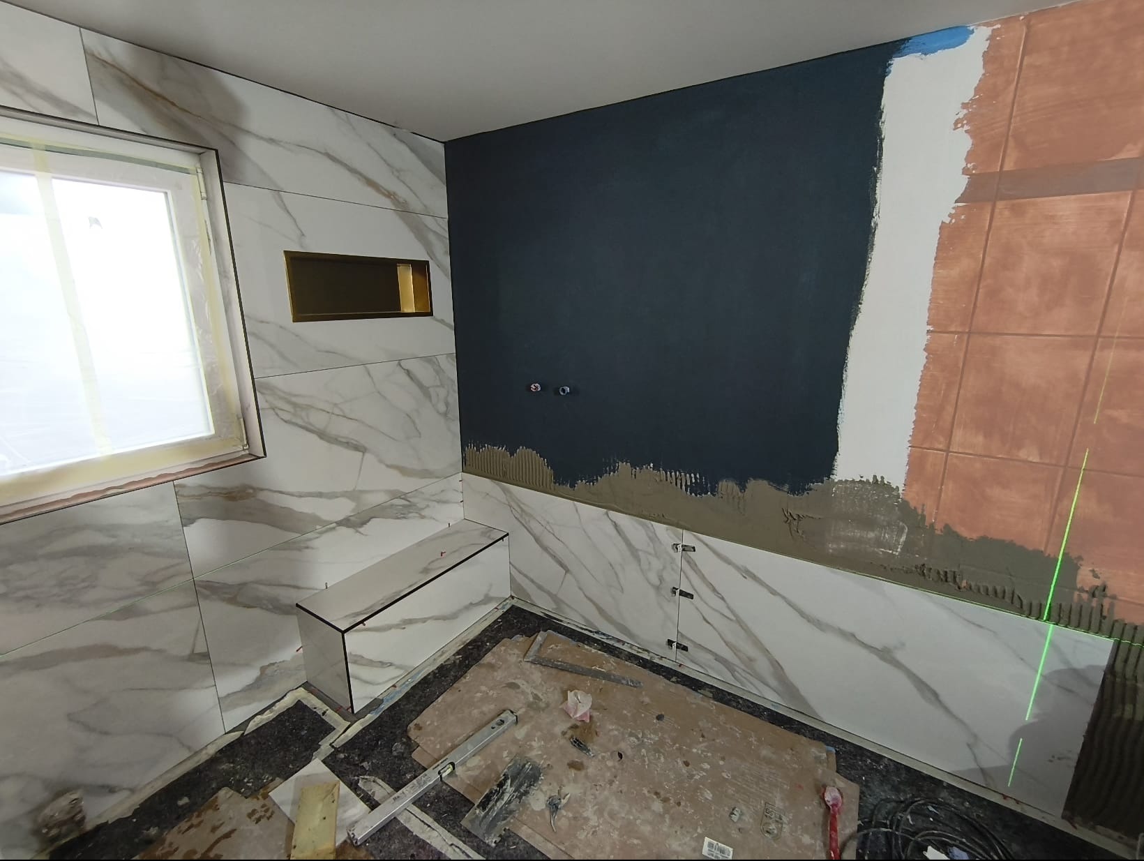 Łazienka w trakcie wykończenia: białe płytki imitujące marmur, wnęka z oświetleniem, fragment ściany z ciemną farbą i laserowy niwelator. Widoczne narzędzia glazurnicze.