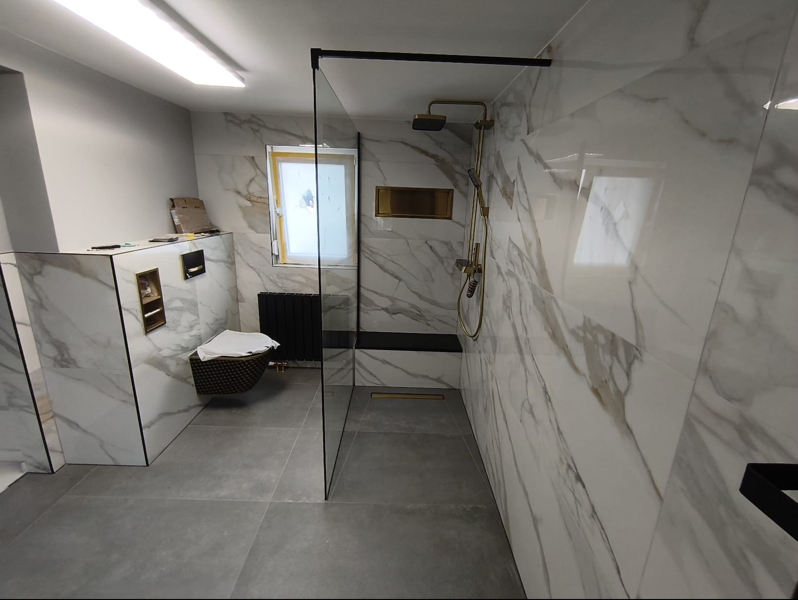 Elegancka łazienka z kabiną typu walk-in, wykończona płytkami imitującymi marmur. Złota armatura, szara podłoga i czarne akcenty tworzą nowoczesny design.