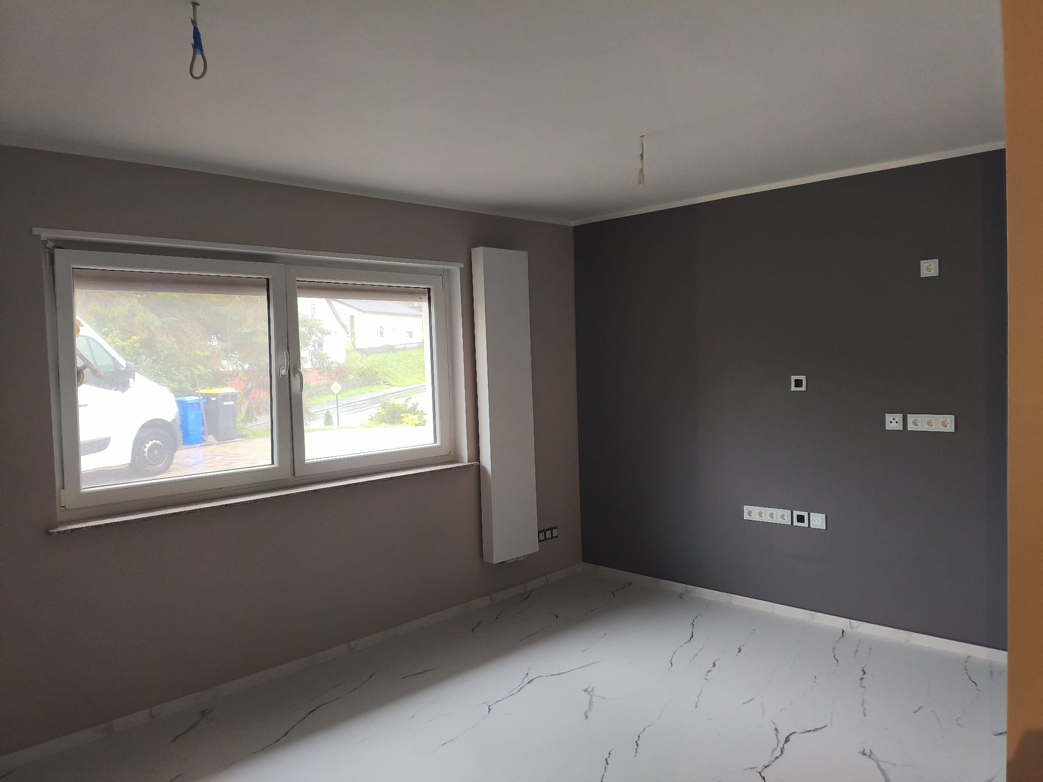 Pokój po malowaniu ścian na szaro i beżowo, z białymi marmurowymi płytkami podłogowymi. Widoczne gniazdka elektryczne i okno z widokiem na zewnątrz.