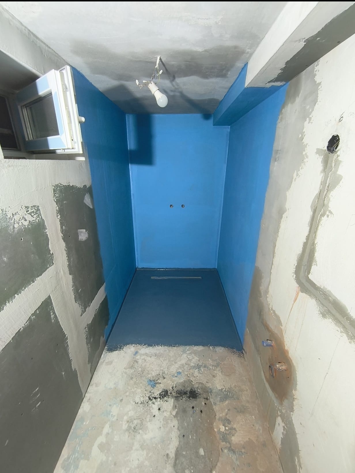 Niebieska hydroizolacja w surowej łazience z widoczną instalacją odpływową i przygotowanymi ścianami do wykończenia. Widoczne surowe ściany i okno.