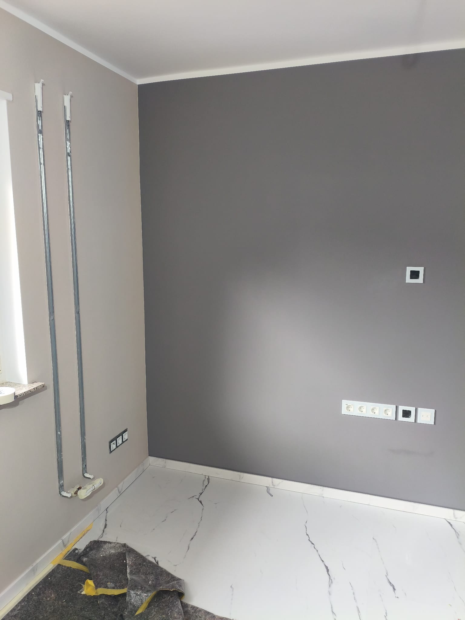 Ściana pomalowana na dwa kolory, z widocznymi puszkami elektrycznymi i rurami grzewczymi. Na podłodze zabezpieczenie. Marmurowe płytki podłogowe.