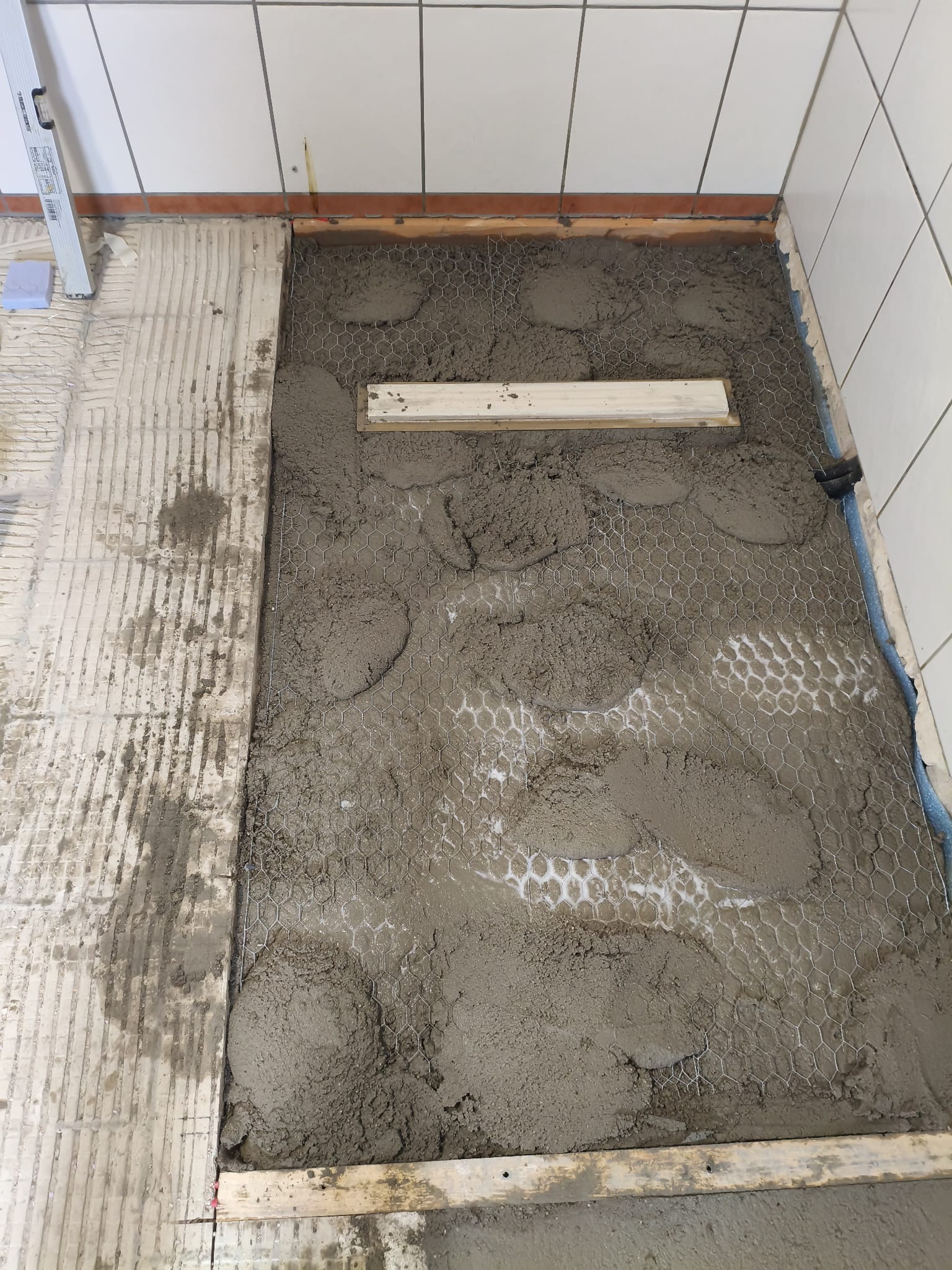 Przygotowanie podłoża w łazience: siatka wzmacniająca zalana częściowo zaprawą, widoczne płytki na ścianach i poziomica. Etap prac przed położeniem płytek.