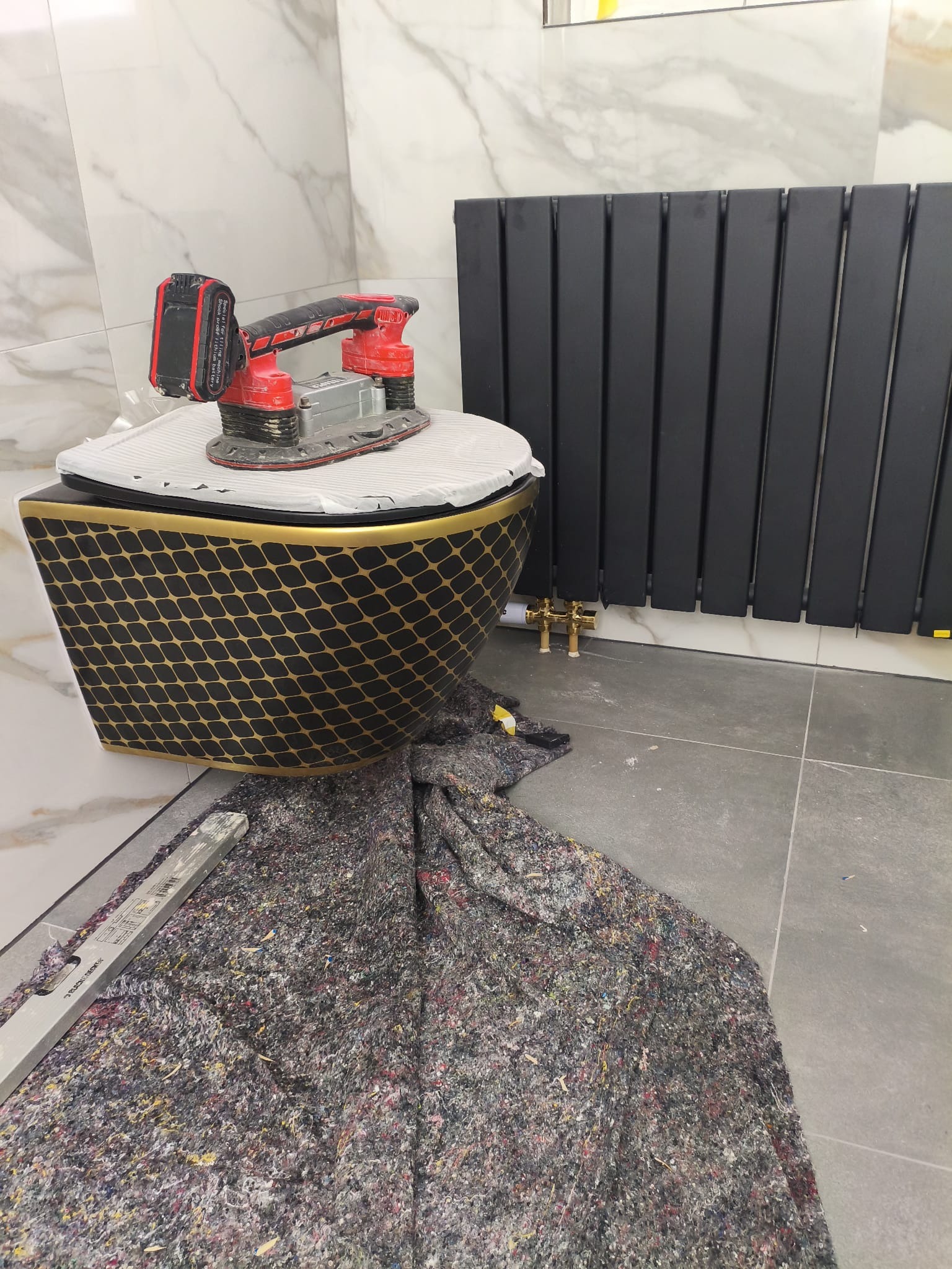 Czarny, dekoracyjny sedes z narzędziem do szlifowania na desce, obok grzejnik drabinkowy i poziomica. Ochronna mata na podłodze w trakcie prac wykończeniowych.