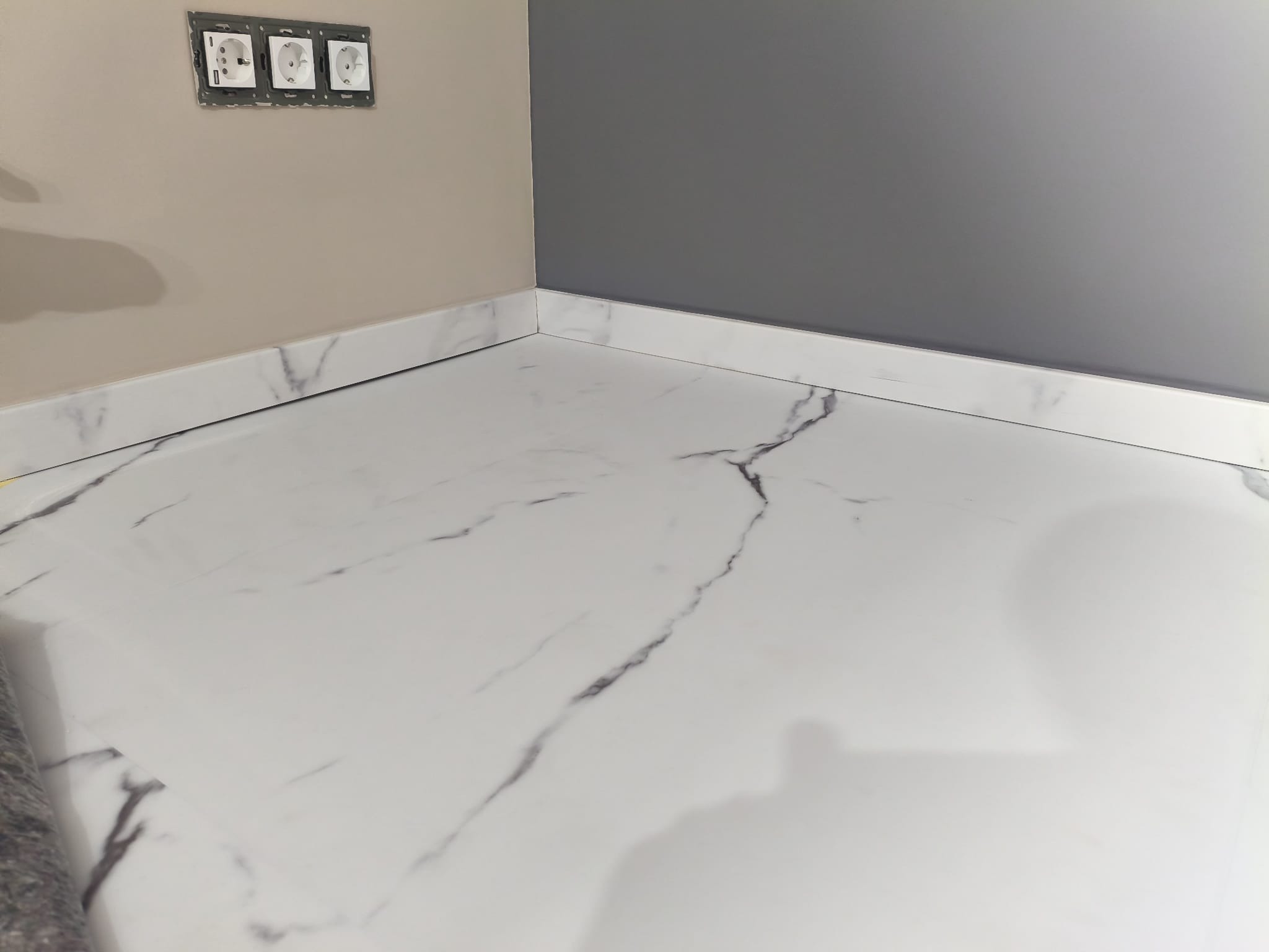 Biała marmurowa podłoga z listwami przypodłogowymi, szara ściana i gniazdka elektryczne. Widok z niskiego kąta, podkreślający wzór marmuru i detale wykończenia.