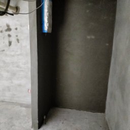 Schulz Krzysztof - Świeżo nałożona, szara gładź szpachlowa na ścianie w rogu pomieszczenia. Widoczny worek z materiałem budowlanym i instalacja elektryczna pod sufitem.