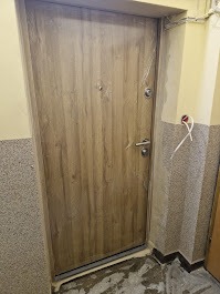 Zamknięte, brązowe drzwi antywłamaniowe z srebrną klamką, zamontowane w korytarzu o szaro-beżowych ścianach. Widoczne przewody elektryczne.