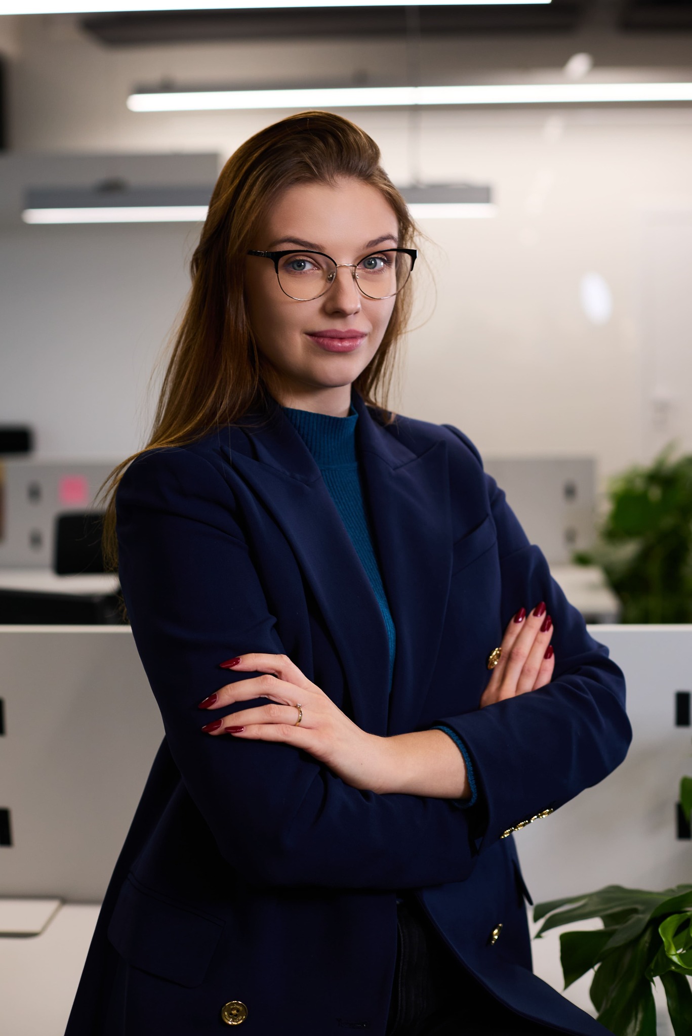 Portret kobiety w okularach i granatowej marynarce, z założonymi rękami na tle biura. Profesjonalny wizerunek, budzący zaufanie.