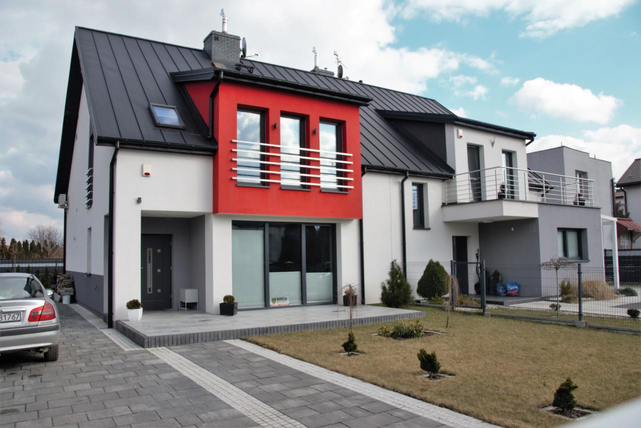 Nowoczesny dom jednorodzinny z czerwoną elewacją, czarnym dachem i szarym brukowanym podjazdem, widok z zewnątrz w słoneczny dzień.