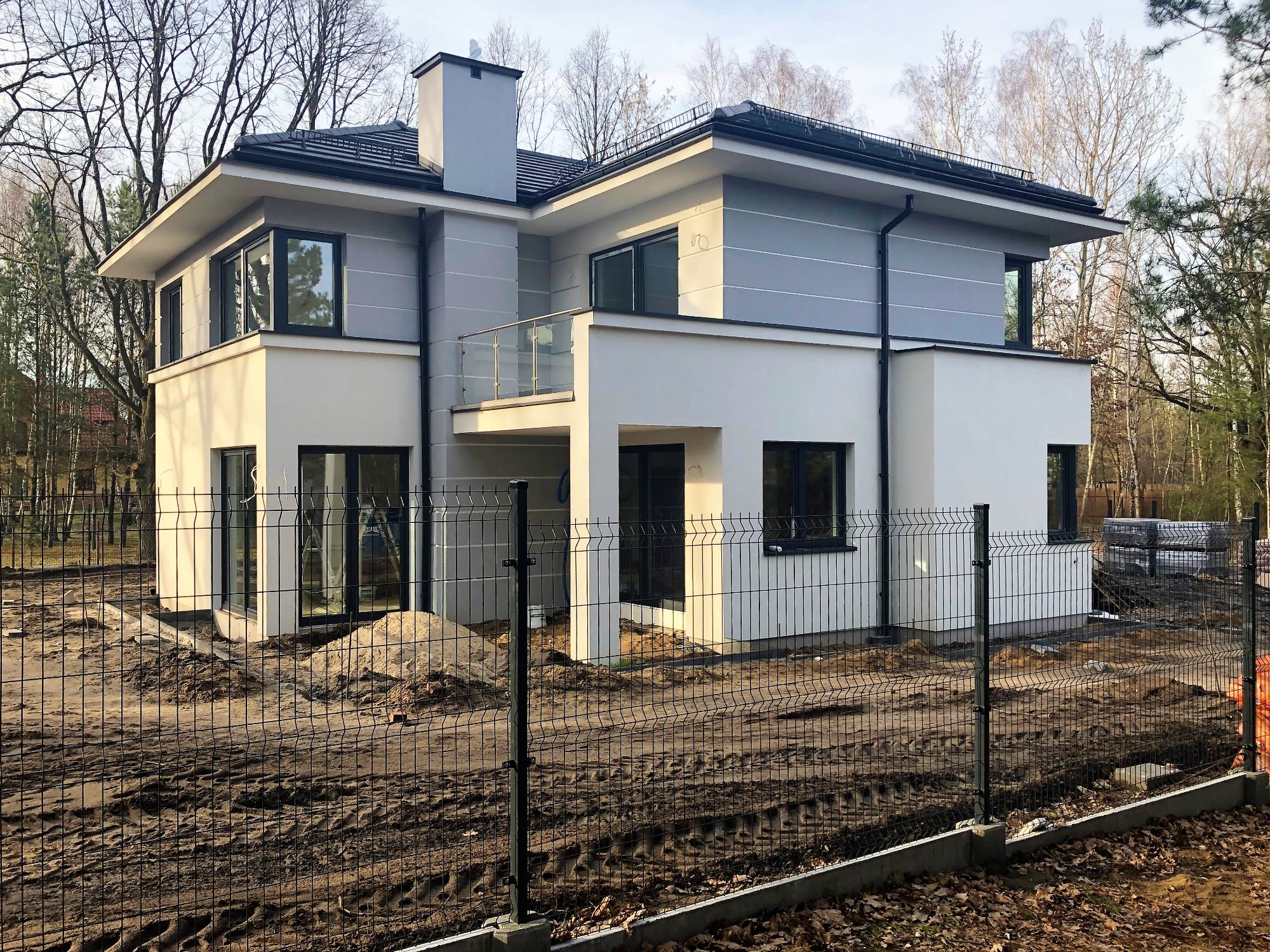 Nowoczesny dwukondygnacyjny dom z płaskim dachem w trakcie budowy, widoczny przez czarne ogrodzenie panelowe, z hałdą piasku obok wejścia.