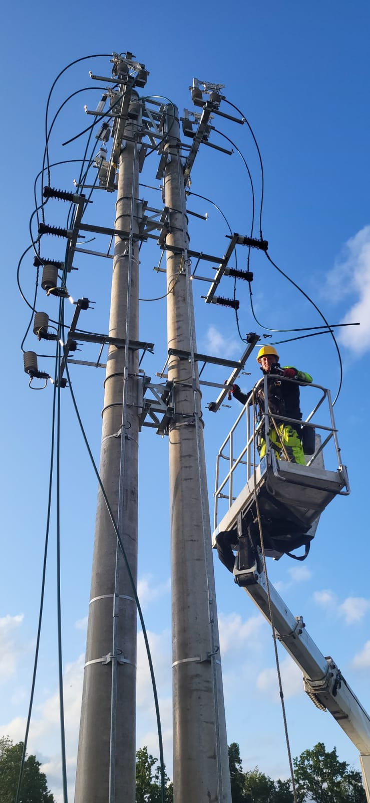 Pracownik w podnośniku koszowym, w kasku, montuje elementy na słupie energetycznym. Widoczne przewody i izolatory na tle błękitnego nieba.