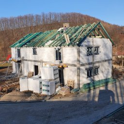 SŁAWOMIR ŁUKOMSKI PROSTY KĄT BUDOWNICTWO - Budowa domu z białych bloczków H+H w trakcie realizacji. Widoczne ściany, okna, więźba dachowa z zielonymi łatami i pracownik na dachu. Na pierwszym planie palety z bloczkami.