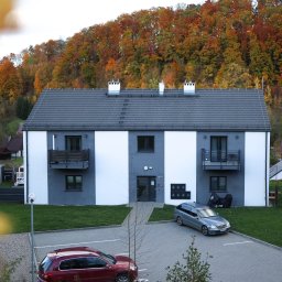 SŁAWOMIR ŁUKOMSKI PROSTY KĄT BUDOWNICTWO - Dwupiętrowy budynek z balkonami w szaro-białej kolorystyce, na tle jesiennego lasu. Na pierwszym planie zaparkowane samochody i kostka brukowa.