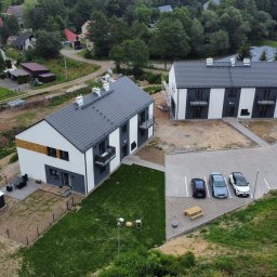 SŁAWOMIR ŁUKOMSKI PROSTY KĄT BUDOWNICTWO - Dwa nowoczesne, dwukondygnacyjne domy z szarymi dachami i białymi elewacjami, widziane z góry. Na parkingu trzy samochody. W tle zieleń i zabudowa.