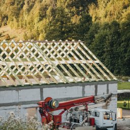 SŁAWOMIR ŁUKOMSKI PROSTY KĄT BUDOWNICTWO - Budowa domu jednorodzinnego: ściany z betonu komórkowego, konstrukcja dachu z drewna, dźwig samochodowy na tle zieleni. Etap wznoszenia konstrukcji.