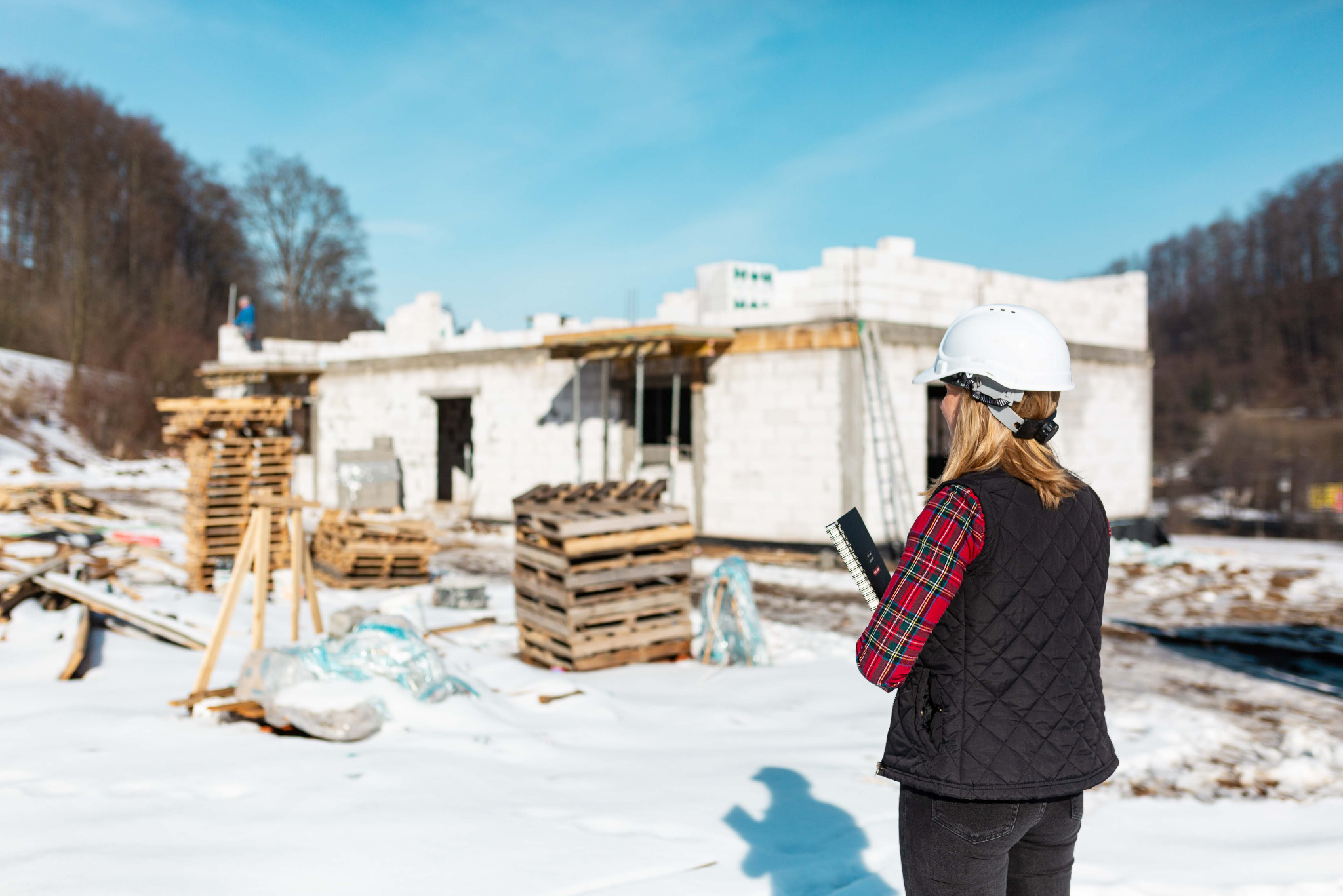 Inspektor w kasku z notatnikiem na tle budowy domu z białych bloczków, zimowy krajobraz. Widoczne palety z materiałami budowlanymi i częściowo wzniesione ściany.