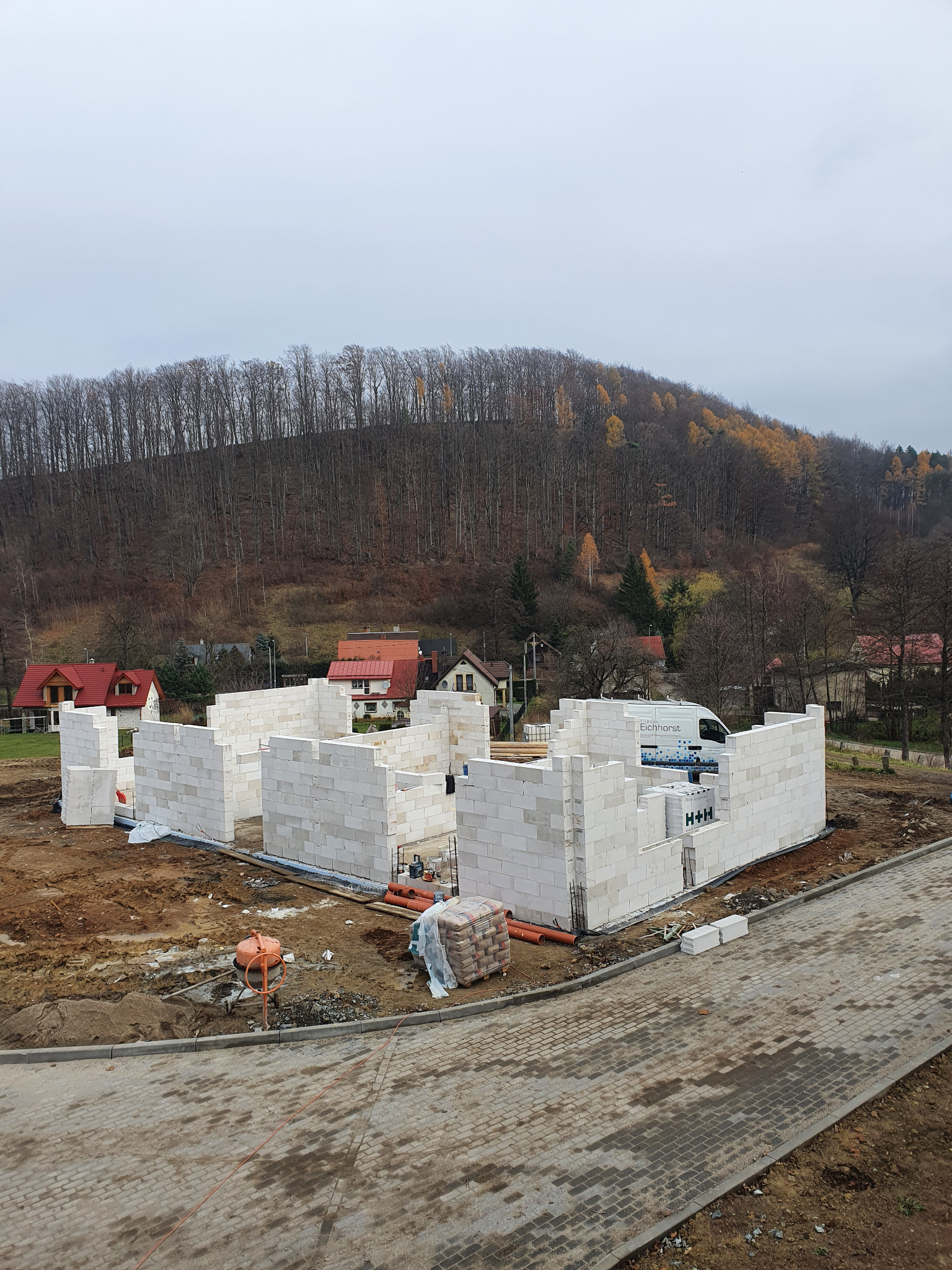 Wznoszenie ścian domu z bloczków H+H na tle zalesionego wzgórza i zabudowań. Widoczna betoniarka i materiały budowlane. Etap budowy w jesiennej scenerii.