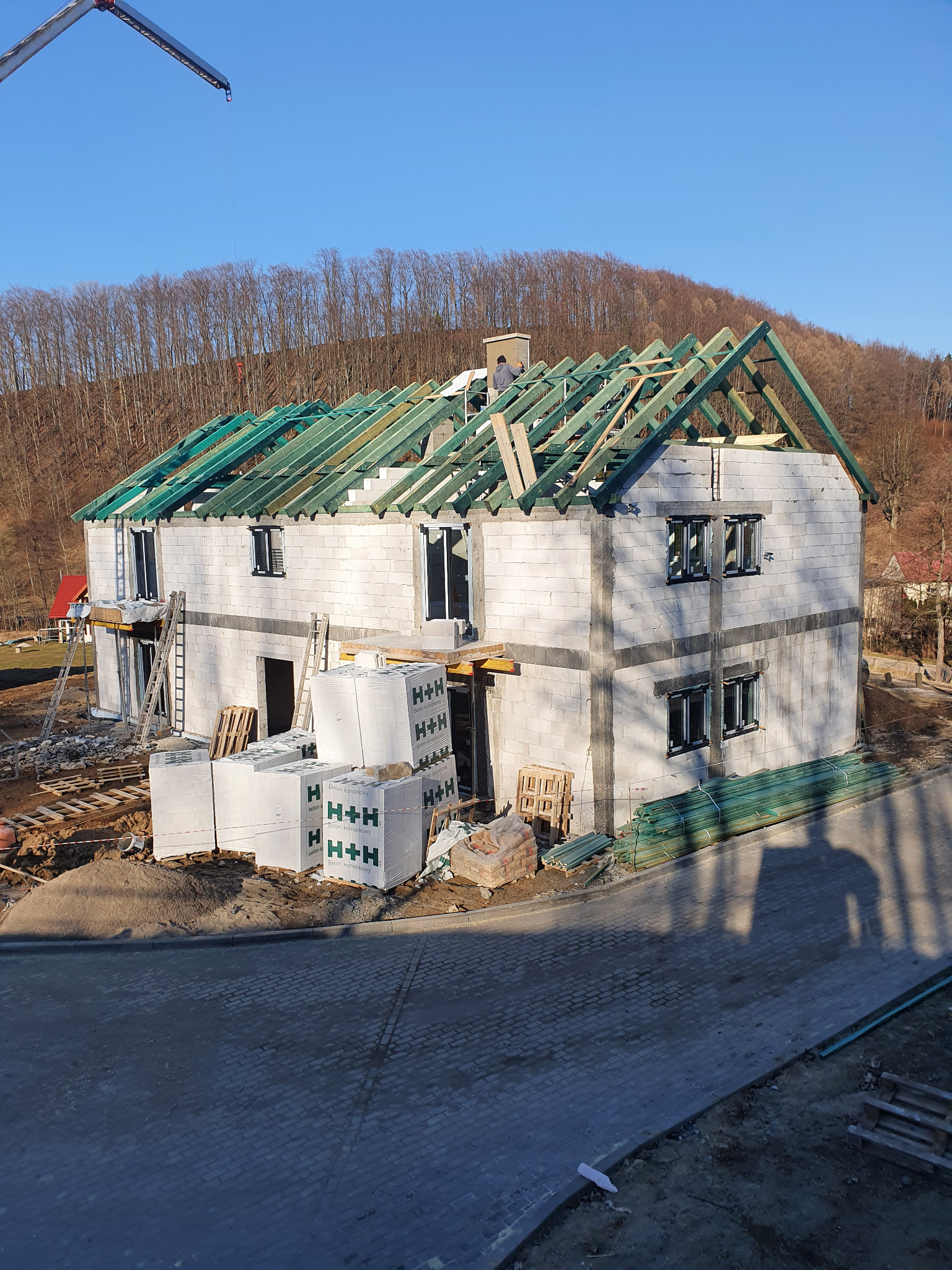 Budowa domu z białych bloczków H+H w trakcie realizacji. Widoczne ściany, okna, więźba dachowa z zielonymi łatami i pracownik na dachu. Na pierwszym planie palety z bloczkami.