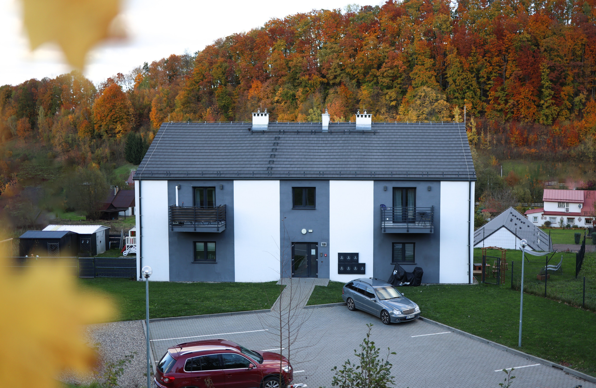 Dwupiętrowy budynek z balkonami w szaro-białej kolorystyce, na tle jesiennego lasu. Na pierwszym planie zaparkowane samochody i kostka brukowa.