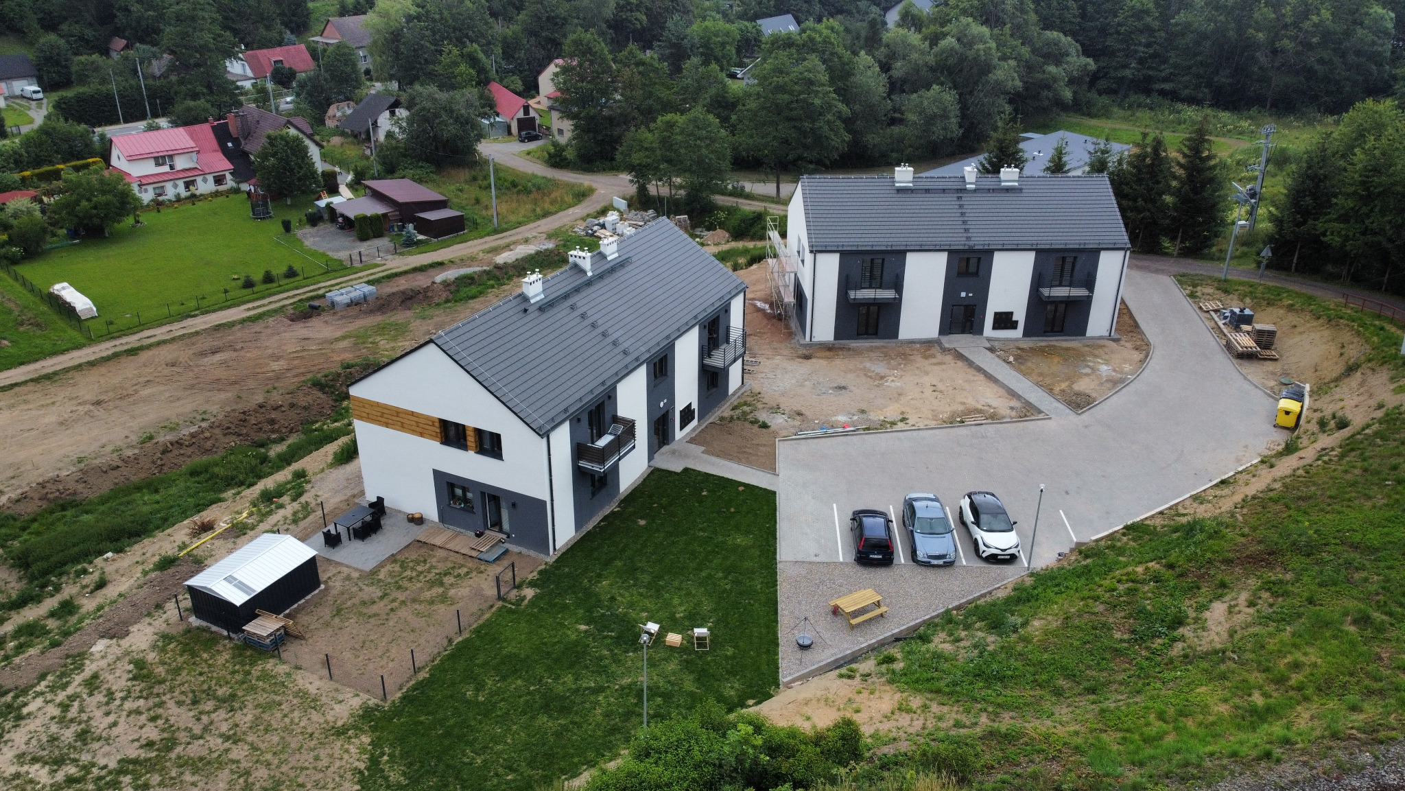Dwa nowoczesne, dwukondygnacyjne domy z szarymi dachami i białymi elewacjami, widziane z góry. Na parkingu trzy samochody. W tle zieleń i zabudowa.