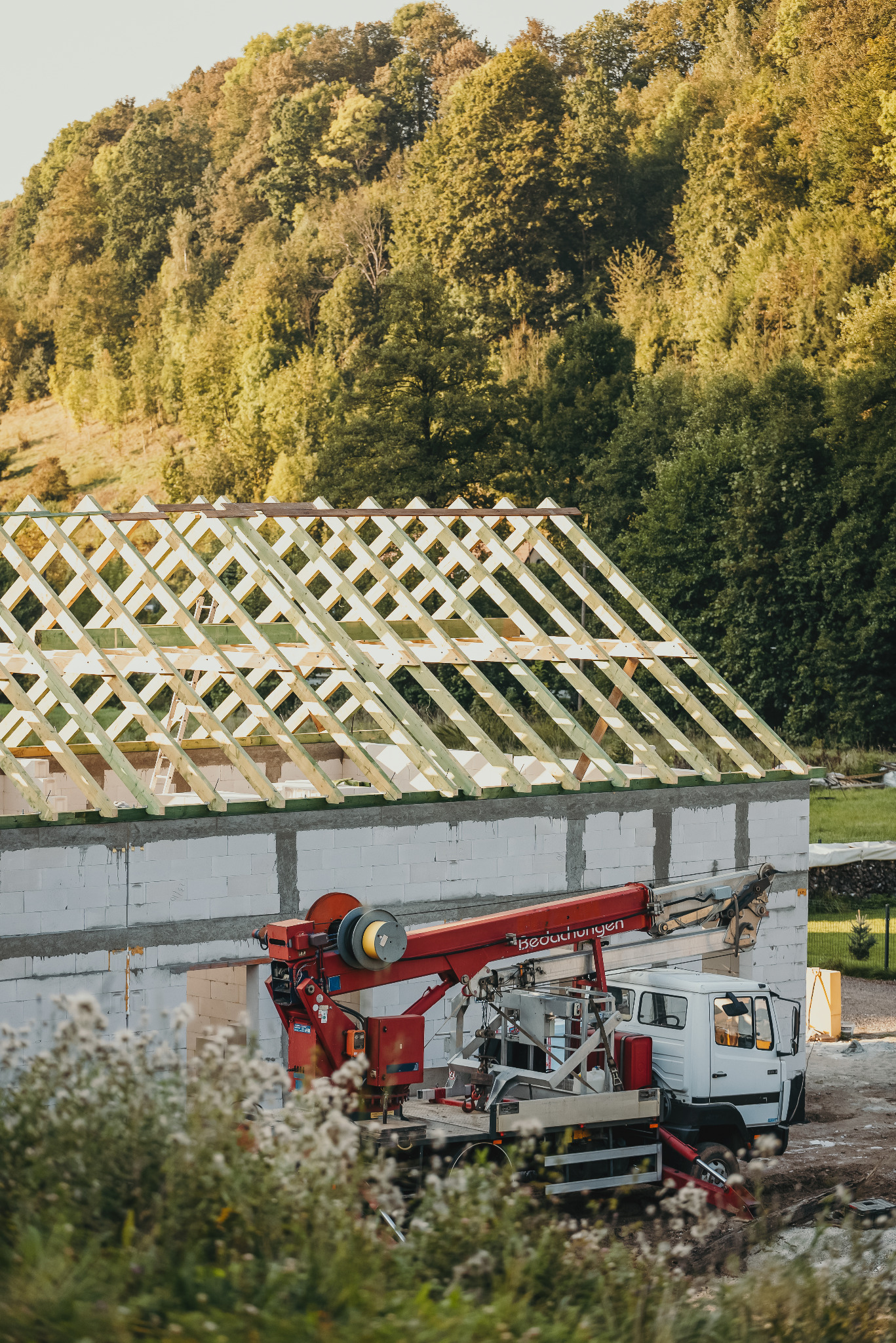 Budowa domu jednorodzinnego: ściany z betonu komórkowego, konstrukcja dachu z drewna, dźwig samochodowy na tle zieleni. Etap wznoszenia konstrukcji.