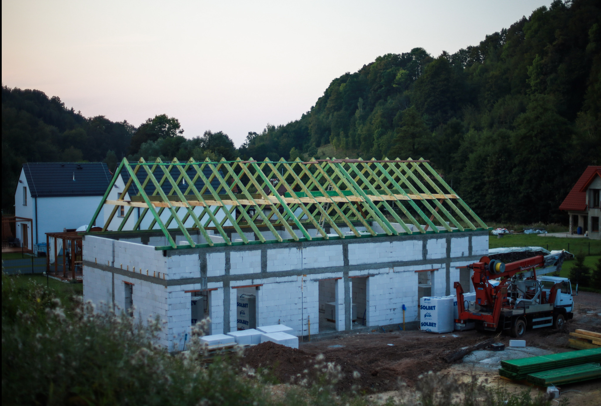 Budowa domu jednorodzinnego z białych bloczków, widoczne ściany i konstrukcja dachu z zielonych łat. Na budowie stoi dźwig samochodowy. W tle zalesione wzgórze.