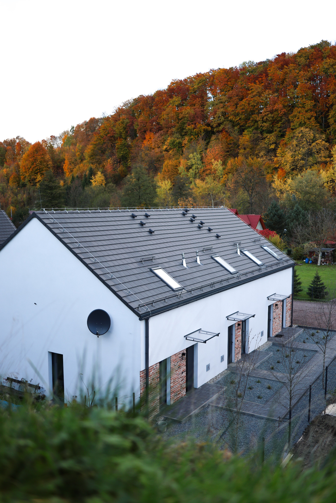 Nowoczesny, biały dom z szarą dachówką i oknami dachowymi, z anteną satelitarną na elewacji. W tle jesienny las i zagospodarowany teren wokół budynku.