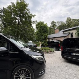 Premium Transfer - Krystian Błotniak - Flota czarnych vanów Mercedes V-klasy na żwirowym podjeździe przed budynkiem. Jeden z vanów z widoczną tablicą rejestracyjną. W tle zieleń drzew i krzewów.