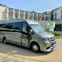 Premium Transfer - Krystian Błotniak - Elegancki, srebrny bus Mercedes-Benz Sprinter z przyciemnianymi szybami, zaparkowany przed hotelem Holiday Inn w słoneczny dzień. Widoczny fragment budynku i logo hotelu.