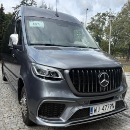 Premium Transfer - Krystian Błotniak - Szary, luksusowy bus Mercedes-Benz Sprinter z chromowanymi lusterkami i charakterystycznym grillem AMG, zaparkowany na brukowanej nawierzchni w słoneczny dzień.