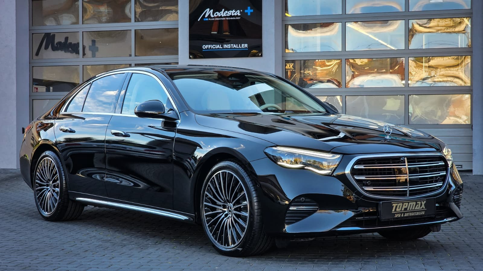 Błyszczący, czarny Mercedes E-Klasa po detailingu, zaparkowany przed salonem z logo Modesta+ i Topmax Spa & Autodetailing. Widoczne odbicia w karoserii.