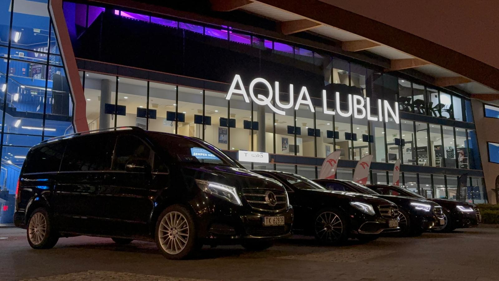 Flota czarnych samochodów Mercedes-Benz, w tym van V-klasy z widoczną tablicą rejestracyjną, zaparkowana przed budynkiem Aqua Lublin nocą, z fioletowym oświetleniem.